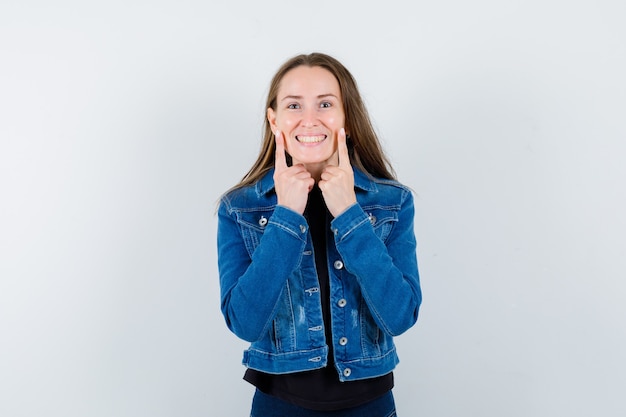 Junge Dame, die Finger auf den Wangen in Bluse, Jacke hält und fröhlich aussieht, Vorderansicht.