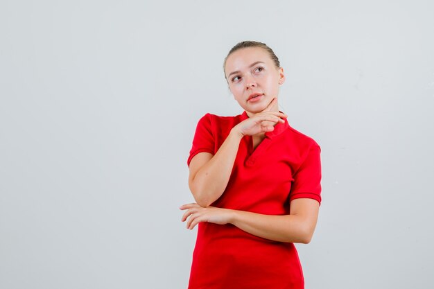 Junge Dame, die Finger auf Kinn im roten T-Shirt stützt und hoffnungsvoll aussieht