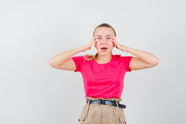 Junge Dame, die Finger auf Schläfen im T-Shirt, in der Hose hält und nett aussieht, Vorderansicht.