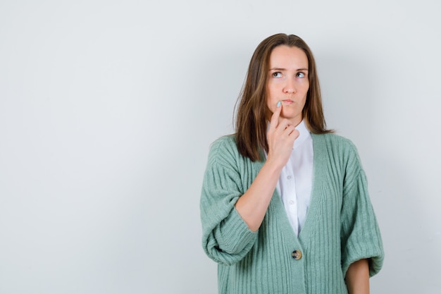 Junge Dame, die Finger nahe Mund im Hemd, in der Strickjacke hält und nachdenklich schaut. Vorderansicht.