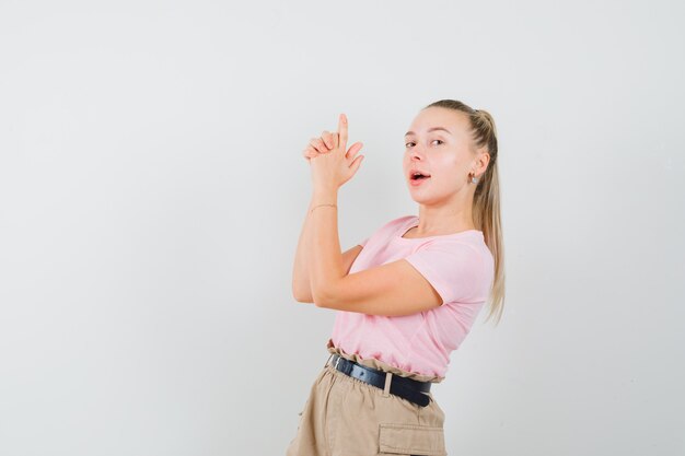 Junge Dame, die Fingerpistolenzeichen im T-Shirt und in der Hose macht und zuversichtlich schaut