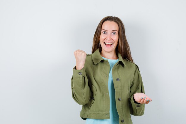 Junge Dame, die geballte Faust in T-Shirt, Jacke hebt und selig aussieht. Vorderansicht.