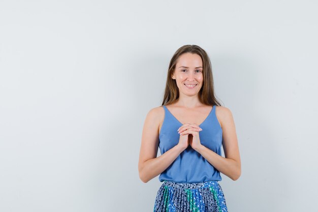 Junge Dame, die gefaltete Finger auf Brust in Unterhemd, Rock hält und hoffnungsvoll aussieht. Vorderansicht.