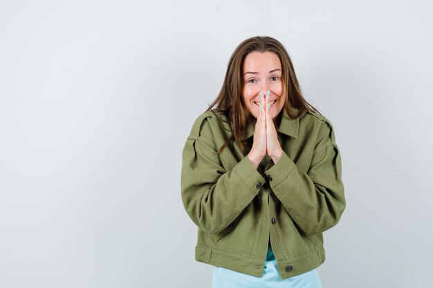 Junge Dame, die gefaltete Hände in flehender Geste in grüner Jacke zeigt und fröhlich aussieht, Vorderansicht.