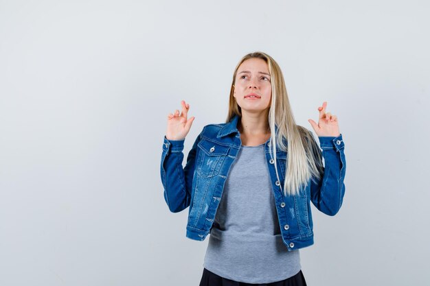 Junge Dame, die gekreuzte Finger in T-Shirt, Jeansjacke, Rock zeigt und nachdenklich aussieht.