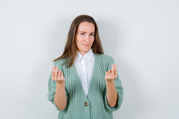 Junge Dame, die Geldgeste in Hemd, Strickjacke macht und zufrieden aussieht. Vorderansicht.