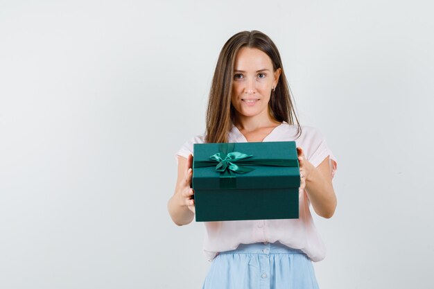 Junge Dame, die Geschenkbox hält und im T-Shirt, Rock, Vorderansicht lächelt.