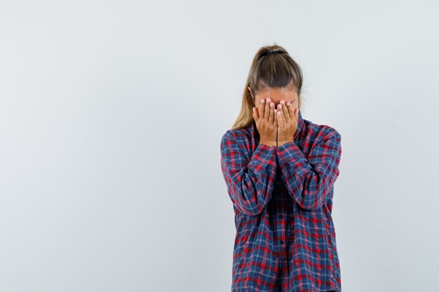 Junge Dame, die Gesicht mit Händen im karierten Hemd bedeckt und deprimiert, Vorderansicht schaut.