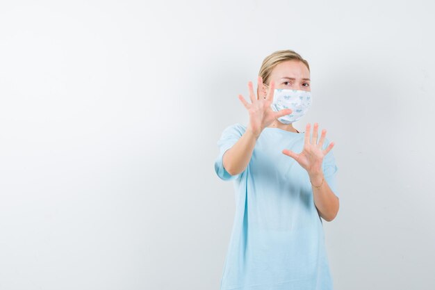 Junge Dame, die Händchen hält, um sich in T-Shirt, Maske und ängstlicher Miene zu verteidigen