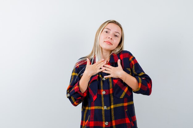 Junge Dame, die Hände auf Brust in kariertem Hemd hält und hoffnungsvoll aussieht. Vorderansicht.