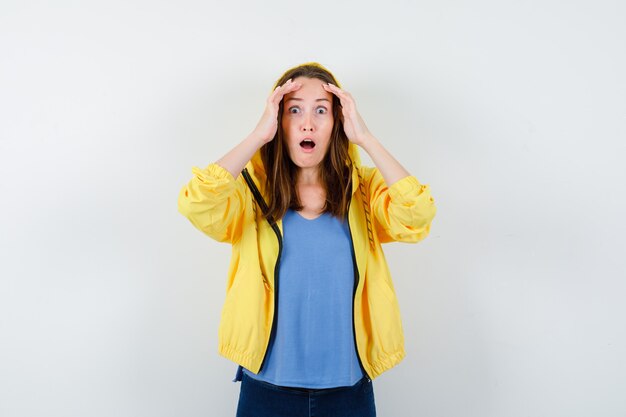 Junge Dame, die Hände auf dem Kopf in T-Shirt, Jacke hält und schockiert schaut, Vorderansicht.