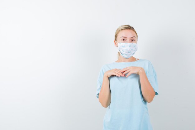 Junge Dame, die Hände auf der Brust in T-Shirt, Maske hält und selbstbewusst aussieht