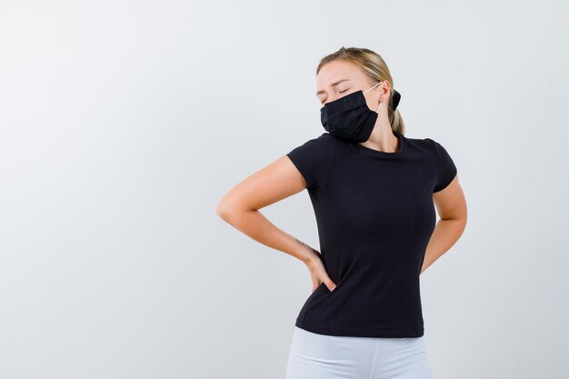 Junge Dame, die Hände auf der Hüfte in T-Shirt, Hose, medizinischer Maske hält und müde aussieht