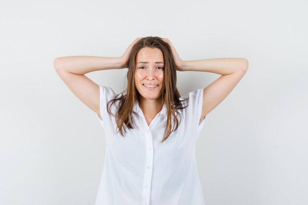 Junge Dame, die Hände auf ihrem Kopf in der weißen Bluse hält und besorgt aussieht