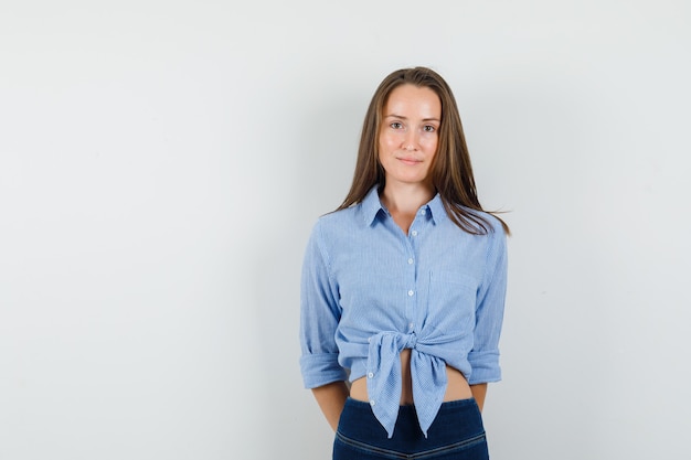 Junge Dame, die Hände auf ihrem Rücken im blauen Hemd, in der Hose hält und optimistisch aussieht