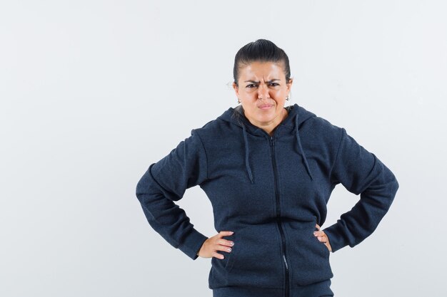 Junge Dame, die Hände auf ihrer Taille hält, während sie ihr Gesicht in der Jacke säuert und unzufrieden aussieht. Vorderansicht.