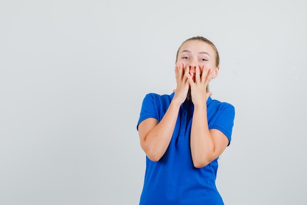 Junge Dame, die Hände auf Mund im blauen T-Shirt hält und glücklich schaut