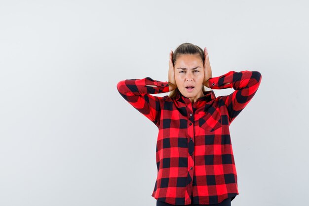 Junge Dame, die Hände auf Ohren im karierten Hemd hält und genervt aussieht