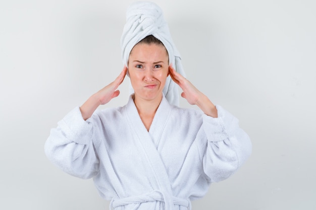 Junge Dame, die Hände auf Ohren im weißen Bademantel, im Handtuch hält und unzufrieden aussieht. Vorderansicht.