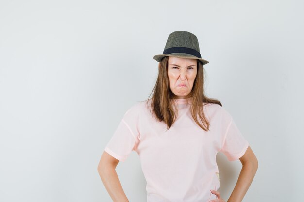 Junge Dame, die Hände auf Taille im rosa T-Shirt Hut hält und boshaft schaut