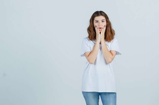 Junge Dame, die Hände auf Wangen im T-Shirt, in den Jeans hält und verwirrt schaut. Vorderansicht.