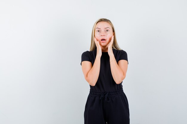 Junge Dame, die Hände auf Wangen im T-Shirt, in der Hose und im ängstlichen Blick von vorne betrachtet.