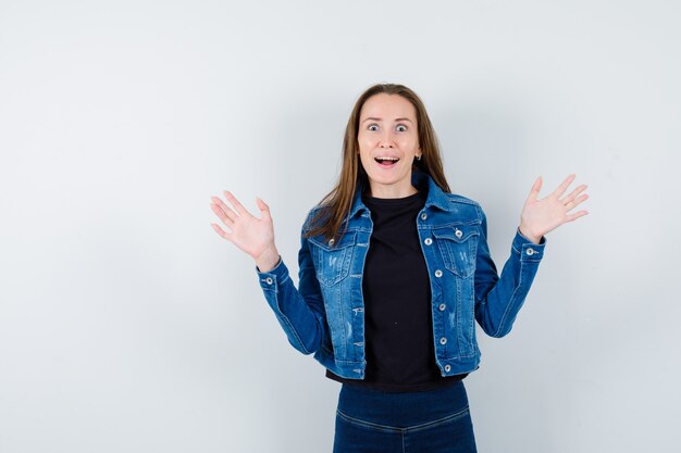 Junge Dame, die Hände hebt, Palmen zeigt, den Mund in Bluse, Jacke öffnet und glücklich aussieht, Vorderansicht.