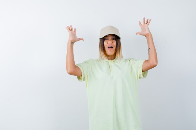 Junge Dame, die Hände in der aggressiven Weise im T-Shirt, in der Mütze und im gestressten Blick von vorne hält.