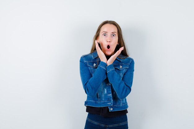 Junge Dame, die Hände in der Nähe des offenen Mundes in der Bluse hält und schockiert schaut. Vorderansicht.