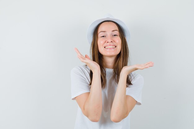 Junge Dame, die Hände nahe Gesicht in weißem T-Shirt Hut hält und niedlich schaut