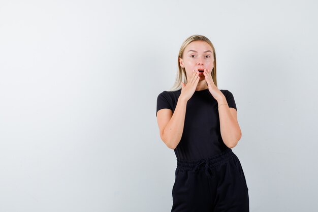 Junge Dame, die Hände nahe Mund in T-Shirt, Hose hält und erstaunt schaut. Vorderansicht.