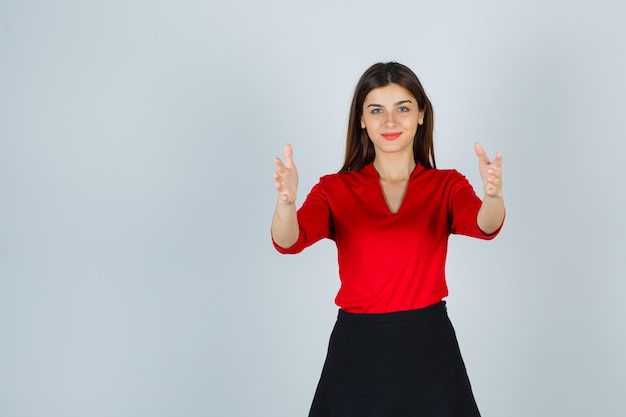 Junge Dame, die Hände streckt, wie Breite in roter Bluse, schwarzem Rock zeigt und glücklich aussieht