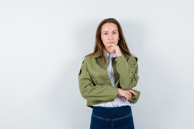 Junge Dame, die Hand am Kinn in Bluse, Jacke hält und vernünftig aussieht, Vorderansicht.