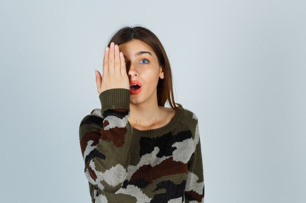 Junge Dame, die Hand auf Auge im Pullover, im Rock hält und verwirrt schaut