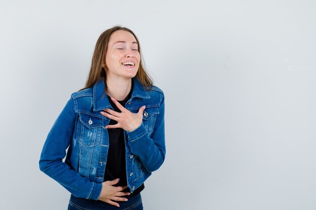 Junge Dame, die Hand auf der Brust in Bluse, Jacke hält und fröhlich aussieht, Vorderansicht.