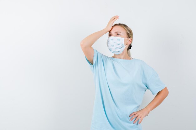 Junge Dame, die Hand auf der Stirn in T-Shirt, Maske hält und nachdenklich aussieht