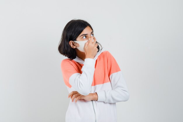 Junge Dame, die Hand auf ihrer Nase im Kapuzenpulli, Gesichtsmaske hält und unzufrieden aussieht.