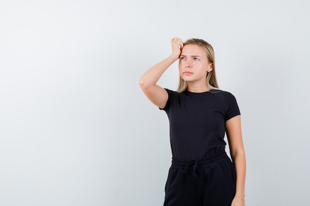 Junge dame, die hand auf kopf im t-shirt, in der hose und nachdenklich aussehend, vorderansicht hält.