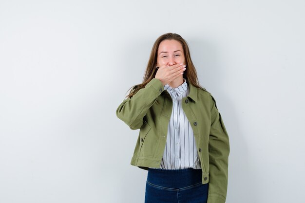Junge Dame, die Hand auf Mund im Hemd, in der Jacke hält und glücklich schaut. Vorderansicht.