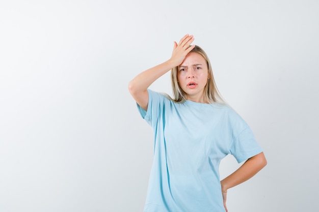 Junge Dame, die Hand auf Stirn hält, während Hand auf Taille im T-Shirt hält und verwirrt, Vorderansicht schaut.