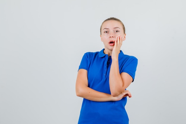 Junge Dame, die Hand auf Wange im blauen T-Shirt hält und überrascht schaut