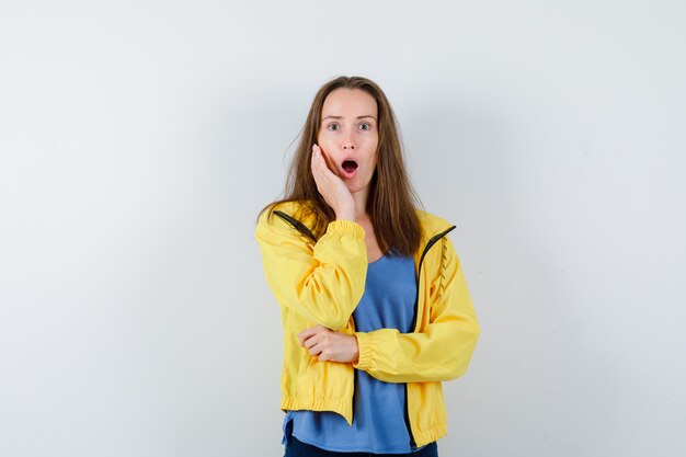 Junge Dame, die Hand auf Wange im T-Shirt, in der Jacke hält und überrascht, Vorderansicht schaut.