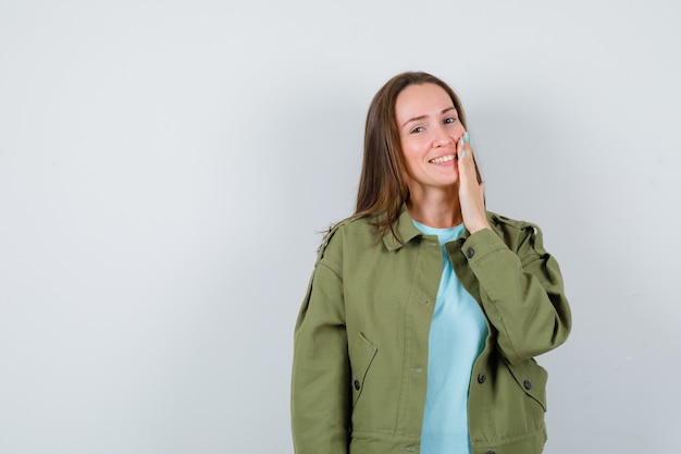 Junge Dame, die Hand auf Wange im T-Shirt, in der Jacke und in der hübschen Vorderansicht hält.