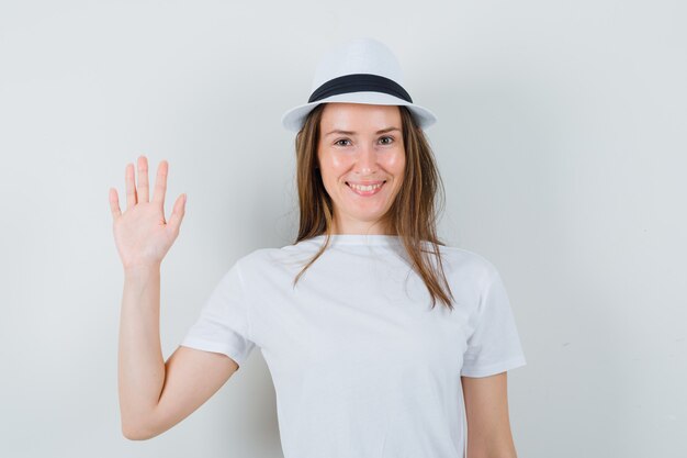 Junge Dame, die Hand winkt, um sich im weißen T-Shirt-Hut zu verabschieden und fröhlich auszusehen
