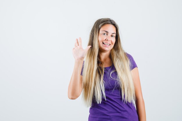 Junge Dame, die Hand zum Begrüßen im violetten T-Shirt winkt und lustige, Vorderansicht schaut.