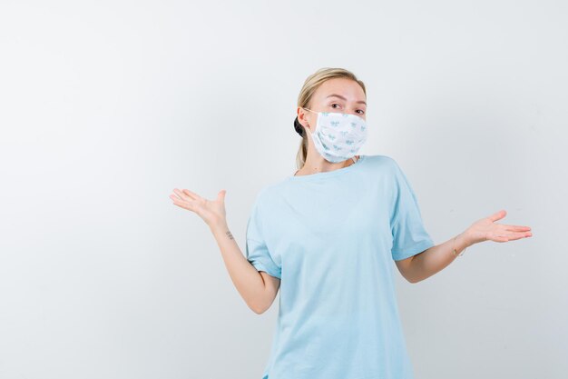 Junge Dame, die Handflächen in T-Shirt, Maske und fröhlichem Gesicht ausbreitet