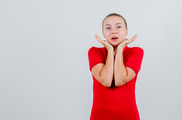 Junge Dame, die Handflächen nahe Kinn im roten T-Shirt hält und amüsiert schaut