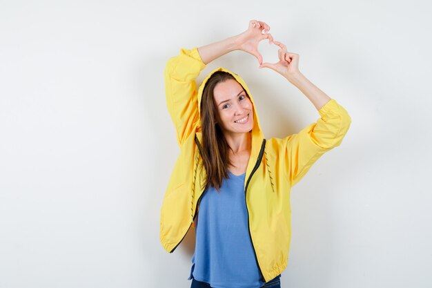 Junge Dame, die Herzgeste im T-Shirt, in der Jacke zeigt und lustig schaut. Vorderansicht.