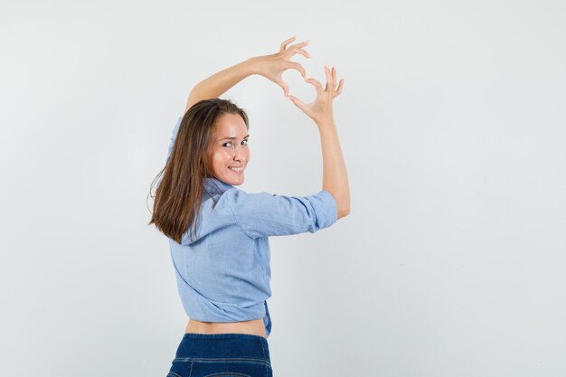 Junge Dame, die Herzgeste zeigt, indem sie im blauen Hemd, in der Hose zurückblickt und glücklich schaut