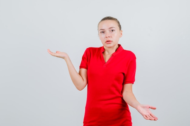 Junge Dame, die hilflose Geste im roten T-Shirt zeigt und verwirrt schaut